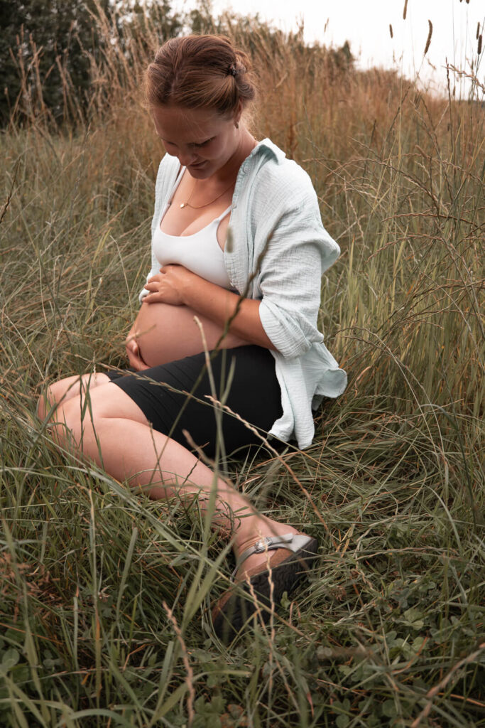 Babybauch Shooting in Hankensbüttel - Frau hält im hohen Gras sitzend liebevoll ihren Babybauch