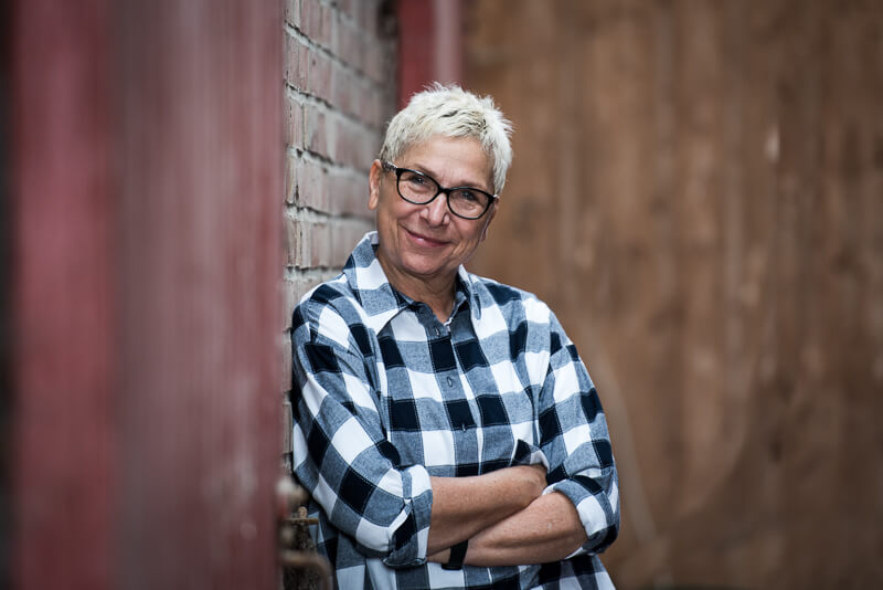 Fotografin aus Hankensbüttel steht lächelnd in einer karrierten Bluse an eine Ziegelwand gelehnt